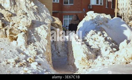 Person walking through narrow path between huge dirty snowdrifts in harsh winter city Stock Photo