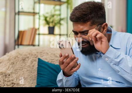 Indian man at home wearing glasses squints reading smartphone text holding screen closer focused. Arabian guy on sofa leans forward eyes narrow tries seeing message shows effort concentration vision. Stock Photo