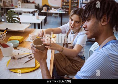 Young woman shaping clay vessel in bright art studio with man observing Stock Photo