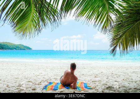 A serene beach scene at Baie Sainte Anne captures a traveler lounging on a colorful towel. Fringed palm leaves sway gently above as the tranquil ocean waves beckon in the distance. Stock Photo