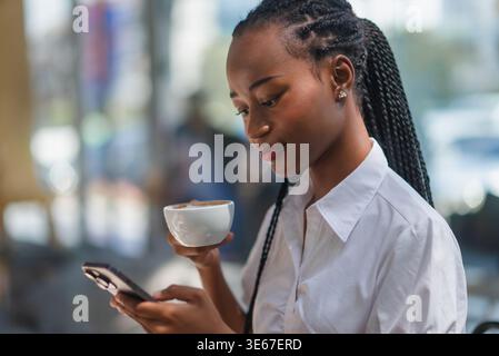 Side view of young Black woman drinking coffee and using smartphone in a cafe. Relaxed lifestyle, mobile technology, online communication, morning rou Stock Photo