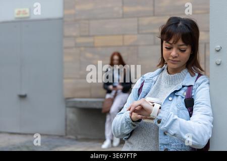 Two women leaning on column by tiled wall on sidewalk, front checking smartwatch, back using phone Stock Photo
