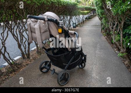 Modern grey and black baby stroller with a orange drink in a bottle holder, parked on a paved pathway between green hedges in a tropical park Stock Photo