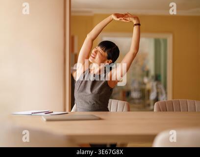Smiling young businesswoman stretching her arms with eyes closed while working at desk in modern office Stock Photo
