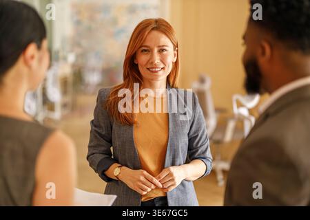 Confident female manager smiling at camera while discussing ideas with professional colleagues in modern office Stock Photo