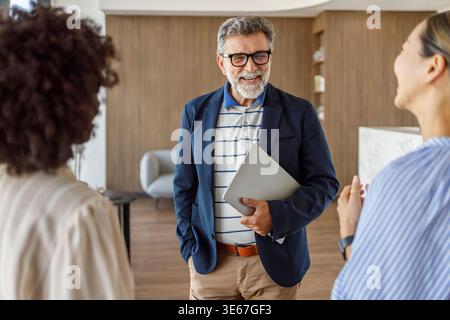 Happy male manager with laptop networking and exchanging ideas with female professionals during collaboration in modern office Stock Photo
