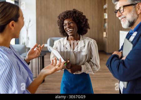 Happy businesswoman holding clipboard and discussing ideas cheerfully with colleagues during collaborative meeting Stock Photo