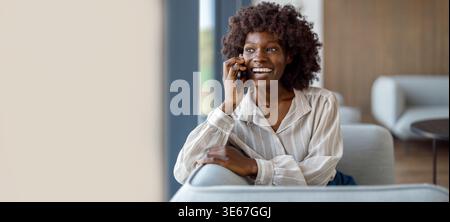 Happy businesswoman with afro hairstyle communicating over smart phone while sitting comfortably on sofa in office Stock Photo