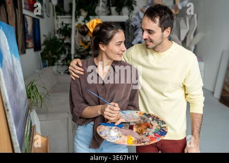 Smiling painters spouses works on painting. Happy woman artist with husband at home studio workshop Stock Photo