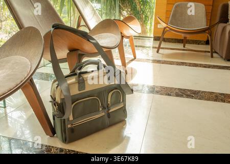 Green professional laptop bag standing on the floor by a wooden chair in a bright lobby with palm trees visible through the window Stock Photo