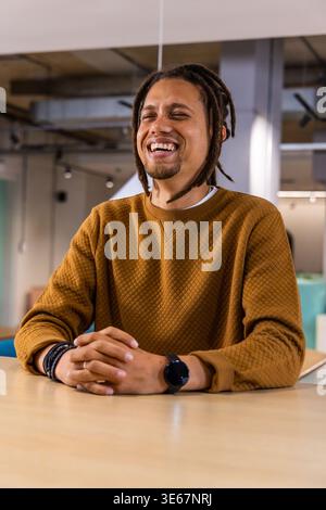 Man in 30s laughing and sitting at office table, wearing brown sweatshirt and black smartwatch Stock Photo