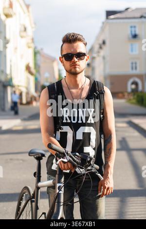 Cool handsome hipster man with stylish sunglasses in a black T-shirt with a bicycle in the city Stock Photo