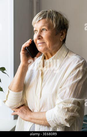 An elderly happy woman enjoys a phone conversation by the window, chatting with loved ones and reflecting on home life Stock Photo