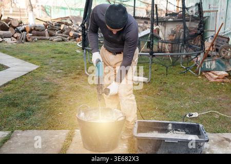 A man stands in a backyard, using an electric mixer to blend cement self-leveling floor in a bucket while wearing gloves and a hat. Tools and logs are Stock Photo