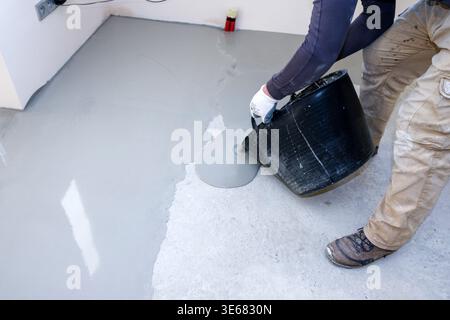 Worker pours cement mixture on concrete self-leveling floor in indoor space during day time for construction project Stock Photo