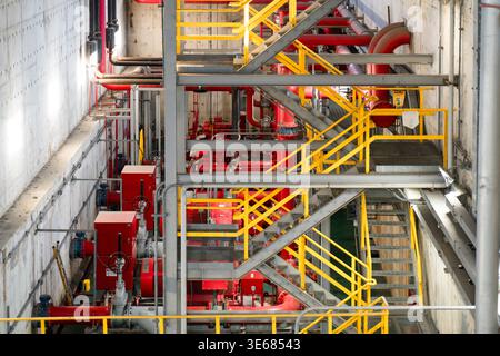 An intricate view of a multi-level industrial facility featuring bright red piping, high-pressure valves, and yellow metal safety stairs. The image hi Stock Photo
