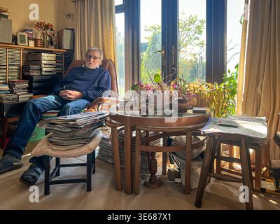 A man sits in a comfy chair in a living room. There is a pile of newspapers on a foot stall. Stock Photo