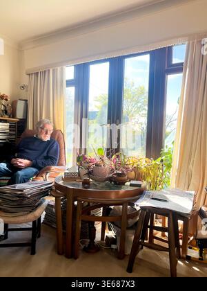 A man sits in a comfy chair in a living room. There is a pile of newspapers on a foot stall. Stock Photo