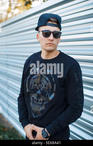 Fashion handsome young hipster man with stylish sunglasses with a baseball cap in a fashionable black pullover on the street near a metal fence Stock Photo