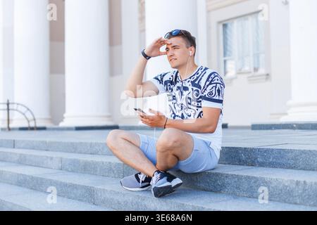 Handsome fashionable young man with sunglasses in a t-shirt with shorts sits on the steps and listens to music in the city Stock Photo