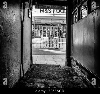 From the end of a dark alleyway onto a street, an old man pedals past on a bicycle. In the background sunlight falls onto the steps of a supermarket. Stock Photo