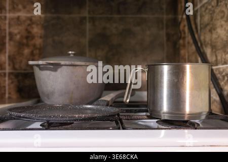 Detailed view of a stainless steel saucepan without lid placed on a gas stove burner. In the background stands a larger cast iron pot with lid and a b Stock Photo