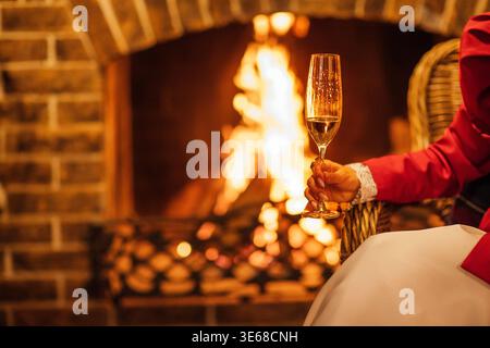Woman in red attire holds a champagne glass near a fireplace, with flames and a brick wall providing a warm and inviting setting Stock Photo
