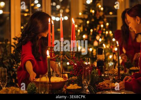 Woman in red sweater places a bowl of food on a beautifully set holiday dinner table with candles and a decorated Christmas tree in the background Stock Photo