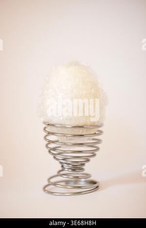 Decorative white egg encased in clear plastic sits atop a spiral metal egg holder, with a soft neutral background enhancing the visual appeal Stock Photo
