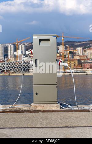 A shore power pedestal with multiple heavy-duty electrical connectors and cables located on a dockside. The background features calm water, high-rise Stock Photo