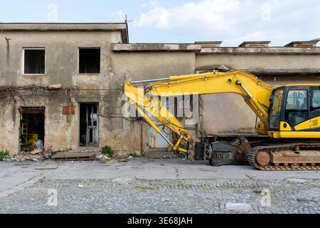 A yellow excavator positioned in front of an old, abandoned building with peeling paint, broken windows, and overgrown vegetation. Construction debris Stock Photo