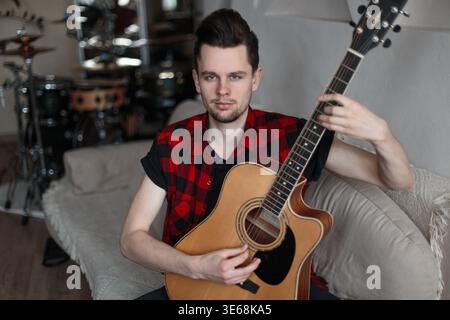 Cool handsome young man with hairstyle in fashion shirt sits on sofa with guitar and practices playing guitar at home Stock Photo