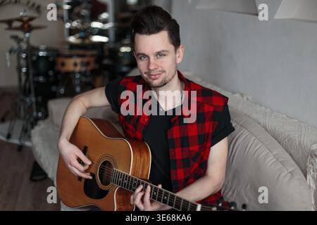 Handsome happy man singer with hairstyle in fashion clothes sitting on sofa with guitar and practicing music playing at home Stock Photo