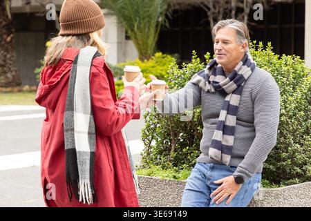 Middle-aged couple exchanging takeaway coffee cups beside low concrete planter, man wearing sweater Stock Photo