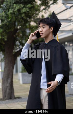 Asian man wearing mortarboard yellow tassel and gown, talking on phone, gesturing on campus path Stock Photo