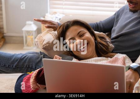 Couple reclining on couch, woman wearing striped sweater holding card while using laptop Stock Photo