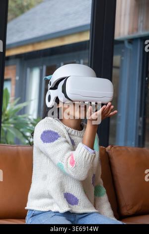 Child wearing white VR headset sitting on brown leather couch at home in knit sweater exploring Stock Photo