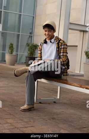 Teenage Asian male student sitting on bench at campus plaza writing in notebook with headphones Stock Photo