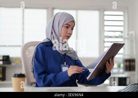 Focused adult Asian woman hijab professional reviewing important documents clipboard office setting business strategy analysis and career development Stock Photo