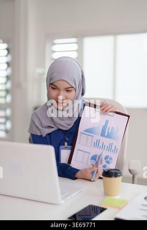 Young Asian Muslim woman professionally discusses financial data analysis charts online Engaged in virtual business meeting from modern office Stock Photo