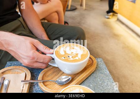 Man hand holding cup of cappuccino with detailed latte art in shape of bear while sitting in cafe served on wooden tray. Close up. Coffee beverage and Stock Photo