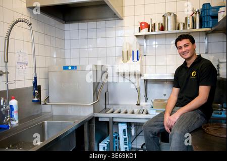 Portrait Young Adult Male working inside a Cafe-Restaurant's Dishwashing Kitchen after school hours to start earning his own income. Rotterdam, Netjerlands. Stock Photo