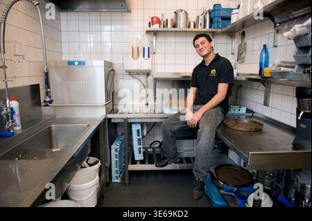 Portrait Young Adult Male working inside a Cafe-Restaurant's Dishwashing Kitchen after school hours to start earning his own income. Rotterdam, Netjerlands. Stock Photo