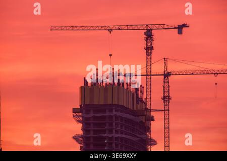 High rise building under construction with tower cranes silhouettes against red sunset sky. Structural framework and lifting equipment in operation Stock Photo