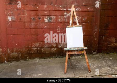 A blank canvas on an easel stands in front of a rustic red brick wall, perfect for artistic expression or presentation Stock Photo