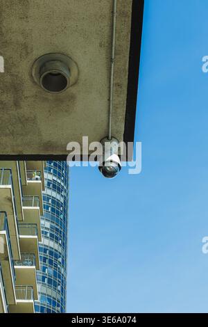 A surveillance camera is installed on the edge of a building while a modern structure with many windows is visible nearby under a clear blue sky in br Stock Photo