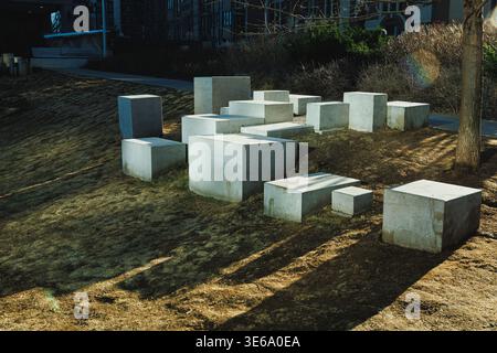 Blocks of concrete are placed on a grassy space beside a walking path in a park. Sunlight creates long shadows on the ground in daylight. Stock Photo