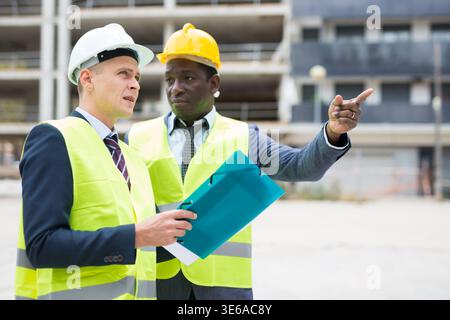 African-american and European men standing on construction site and talking. One of them holding folder with documentation, another one showing Stock Photo