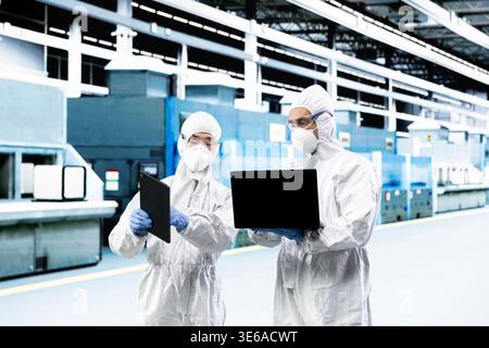 Team of technicians in PCB factory cleanroom monitoring production lines equipment telemetry. Teamworking colleagues wearing safety gear using devices to document microchip yield variations Stock Photo