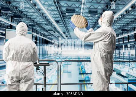 Cleanroom technicians inspecting silicon wafer on elevated smart factory platform. Scientists evaluating semiconductor substrates produced by automated machinery in industrial 4.0 manufacturing plant Stock Photo